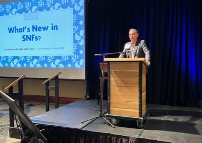 Oregon Geriatrics Society 2023 Conference - an attendee presenting at a podium on a raised platform with a dark blue velvet curtain behind them - to the left is a projector screen with the title card "What's New in SNFs?"