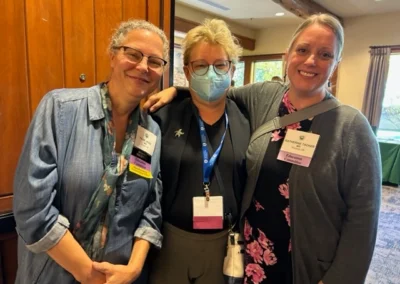 Oregon Geriatrics Society 2023 Conference - three attendees smile for a photo - one has her arm around another's shoulder - rustic lodge