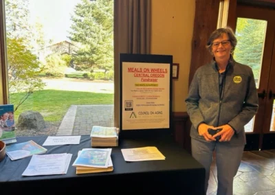Oregon Geriatrics Society 2023 Conference - an attendee poses for a photo next to a table with pamphlets and a sign reading "Meals on Wheels Central Oregon Fundraiser" - a window behind the table shows trees and a stone walkway outside