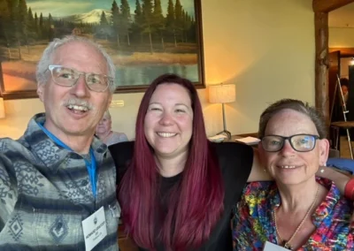 Oregon Geriatrics Society 2023 Conference - three attendees smile for a photo in a room with cream-colored walls. A painting of a river, a log cabin, pine trees and Mt. St. Helens is hung on the wall behind them.