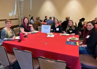 Oregon Geriatrics Society 2023 Conference - five attendees seated at a rectangular table with a bright red tablecloth eating dinner - a reservation card reading "Acute Care Consult Service" is in the center - cream-colored walls, other attendees in background