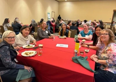 Oregon Geriatrics Society 2023 Conference - seven attendees seated at a rectangular table with a bright red tablecloth in the foreground of a packed conference hall - rustic lodge