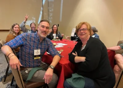 Oregon Geriatrics Society 2023 Conference - six attendees seated at a rectangular table with a bright red tablecloth eating dinner - one of them is throwing up a peace sign - rustic lodge