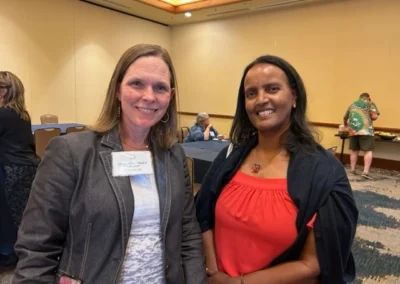 Oregon Geriatrics Society 2023 Conference - two attendees pose for a photo in a room with pale yellow walls and a patterned floor - a few attendees are in the background seated at tables and eating from the buffet