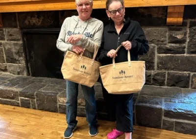 Oregon Geriatrics Society 2024 Conference - two attendees posing with Sunriver Resort branded tote bags in front of a stone brick fireplace with a long wooden mantle.