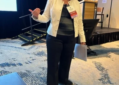 Oregon Geriatrics Society 2024 Conference - a woman standing adjacent to the stage where two men are behind the podium, blue curtain in background, projector screen to her back right. She is speaking to someone out of frame and holding a sheet of paper.