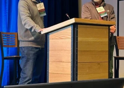 Oregon Geriatrics Society 2024 Conference - two gentleman standing behind a podium, one of whom is speaking and addressing the room. Blue curtain in background