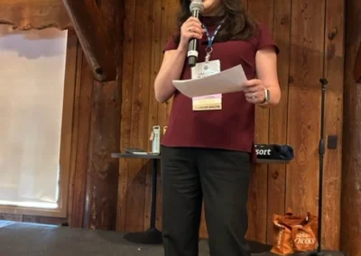 Oregon Geriatrics Society 2024 Conference - an attendee holding a sheet of paper speaks into a microphone onstage in a rustic lodge with a projector screen in the background to the left