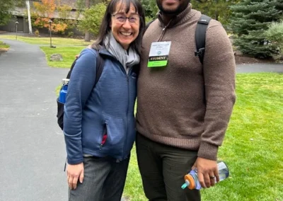 Oregon Geriatrics Society 2025 Conference - a man and woman standing close together outside on a concrete walkway surrounded by trees. The man is carrying a water bottle and wearing a "Student" nametag