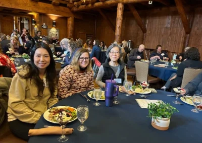 Oregon Geriatrics Society 2025 Conference - three attendees sitting at a table eating dinner in foreground, numerous attendees in background - blue tablecloth, lovely centerpiece, rustic lodge