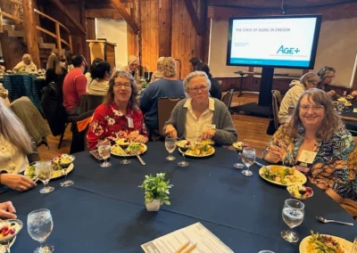 Oregon Geriatrics Society 2025 Conference - three attendees eating dinner at a round table - blue tablecloth, lovely centerpiece - rustic lodge - in the background is a screen showing a page of a presentation, "The State of Aging in Oregon", Age+