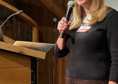 Oregon Geriatrics Society 2025 Conference - a woman speaking into a microphone held in her right hand, standing to the right of a nearby podium - rustic lodge