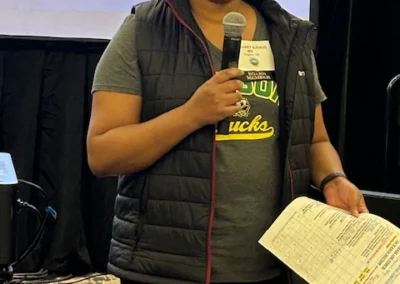 Oregon Geriatrics Society 2025 Conference - a member of the board speaking into a microphone held in her right hand. In her left hand is a sheet with marks and highlighted text. A projector screen displays a logo behind her head.