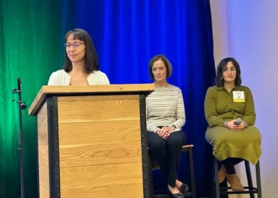 Oregon Geriatrics Society 2025 Conference - two attendees seated in front of an emerald curtain with a blue spotlight coming down from above. In the foreground, a third attendee prepares to speak at a podium.