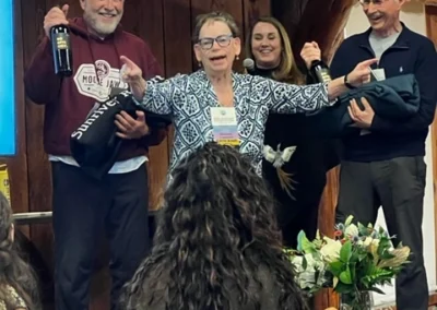 Oregon Geriatrics Society 2024 Conference - an attendee points cheerfully with both hands at the crowd as three other attendees stand behind her smiling and holding up bottles of wine. Two seated attendees look on in the foreground.